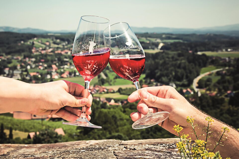 Zwei Gläser mit roséfarbenem Wein werden zu einem Prosit angehoben. Im Hintergrund ist eine malerische Landschaft mit grünen Feldern und Dörfern zu sehen. | © Schilcherland Steiermark – Lupi Spuma
