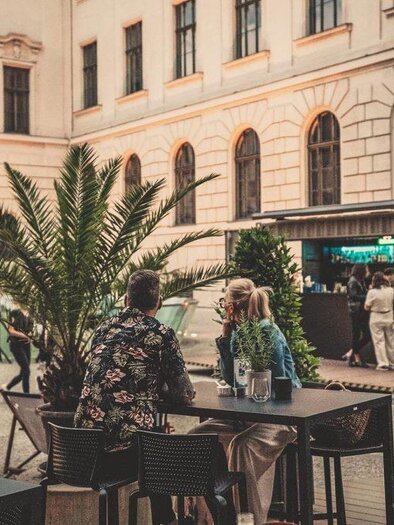 Ein lebhaftes café im Freien mit Palmen und Tischen. Menschen genießen die Atmosphäre in einer städtischen Umgebung. | © CIS