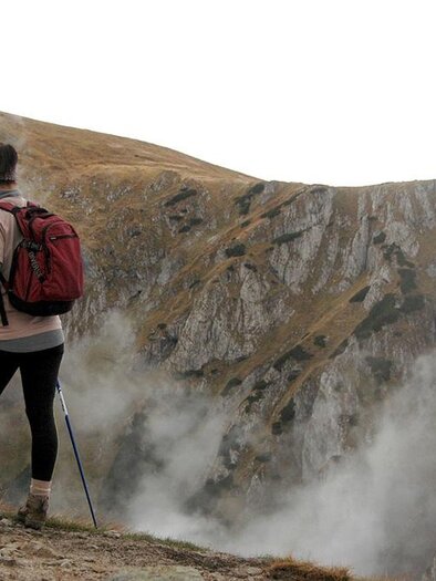 Eine Wanderin steht an einer felsigen Klippe und blickt in die Ferne. Der Himmel ist bewölkt und Nebel umhüllt die umliegenden Berge. | ©  Pexels