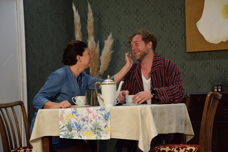 Ein Paar sitzt an einem Tisch und genießt eine gemütliche Zeit zusammen. Die Frau berührt sanft das Gesicht des Mannes, während sie eine Tasse Kaffee trinken. | © Theatergruppe Vogau
