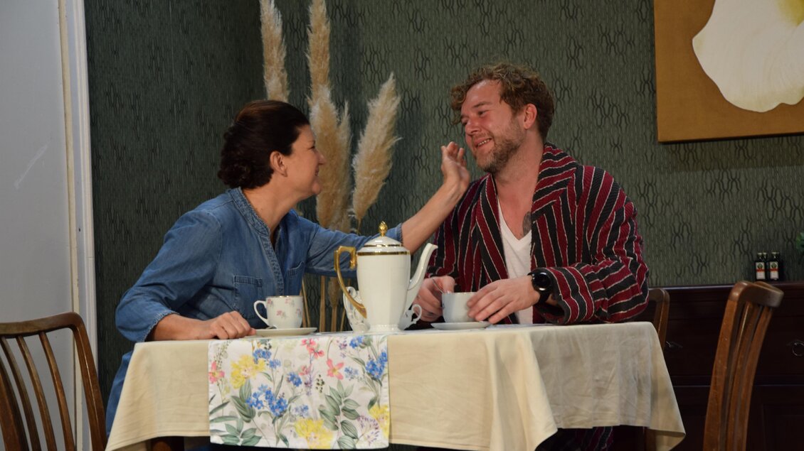 Ein Paar sitzt an einem Tisch und genießt eine gemütliche Zeit zusammen. Die Frau berührt sanft das Gesicht des Mannes, während sie eine Tasse Kaffee trinken. | © Theatergruppe Vogau
