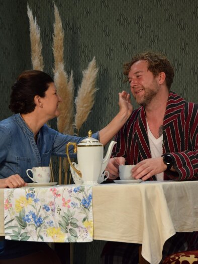 Ein Paar sitzt an einem Tisch und genießt eine gemütliche Zeit zusammen. Die Frau berührt sanft das Gesicht des Mannes, während sie eine Tasse Kaffee trinken. | © Theatergruppe Vogau