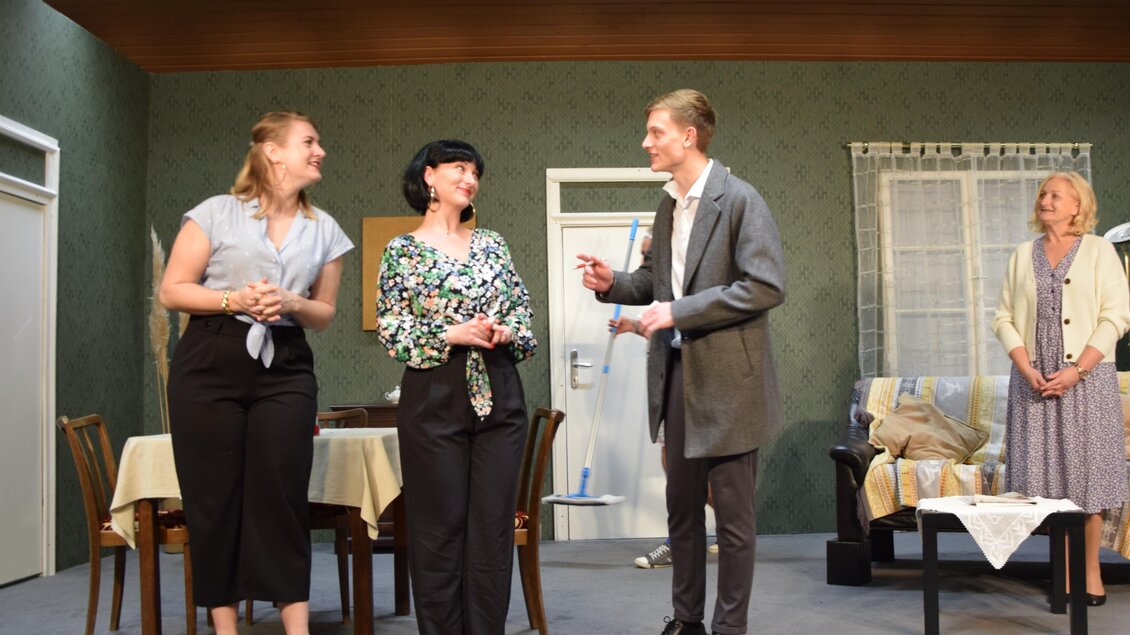 Eine Bühneninszenierung mit vier Schauspielern in förmlicher Kleidung. Sie stehen in einem Wohnzimmer mit einem Tisch und Stühlen im Hintergrund. | © Theatergruppe Vogau