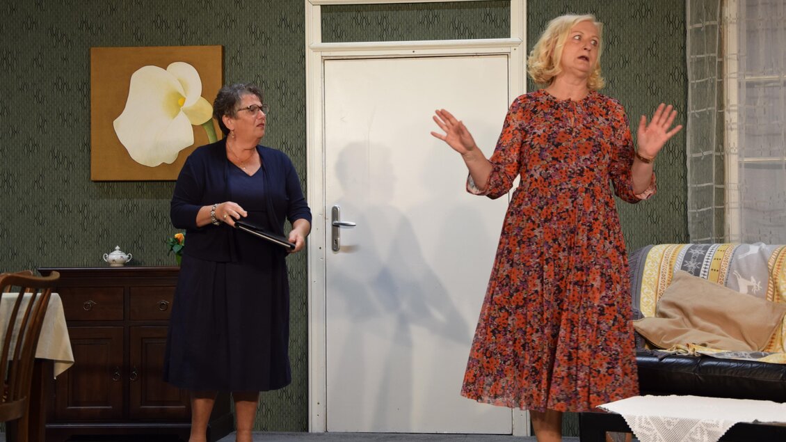Zwei Frauen in einem Wohnzimmer, eine trägt ein florales Kleid und gestikuliert, während die andere mit einem Objekt in der Hand zuschaut. Die Wände sind grün dekoriert und ein Bild einer Blume hängt an der Wand. | © Theatergruppe Vogau