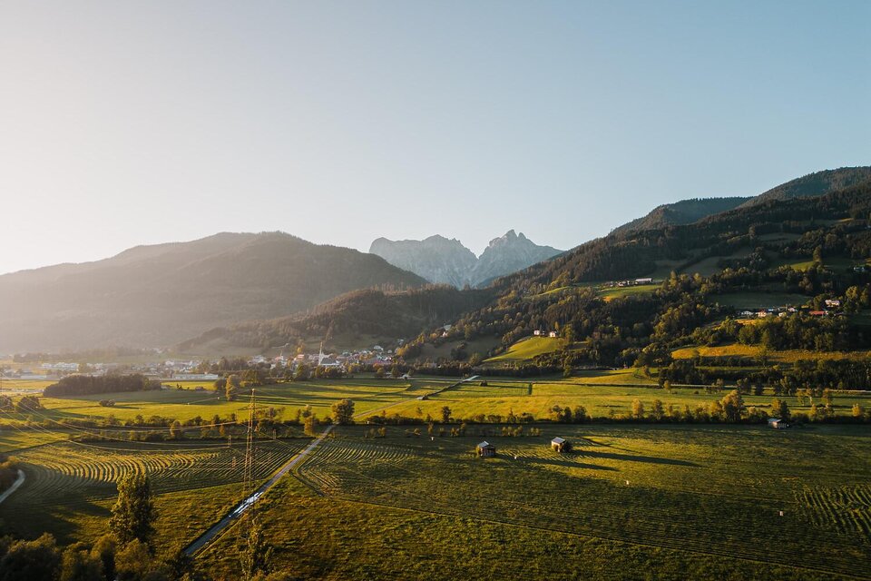 Eine malerische Landschaft mit sanften Hügeln und schneebedeckten Bergen im Hintergrund. Die Wiesen sind grün und das Licht der Abendsonne erzeugt eine warme Atmosphäre.