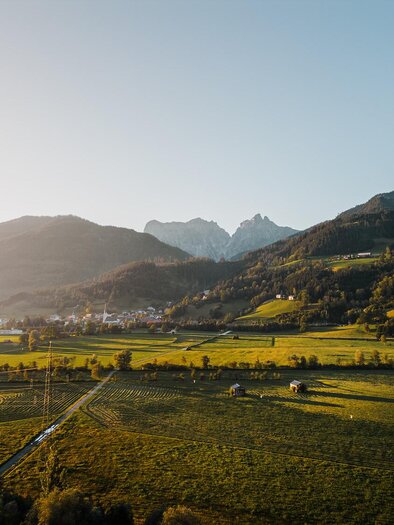 Eine malerische Landschaft mit sanften Hügeln und schneebedeckten Bergen im Hintergrund. Die Wiesen sind grün und das Licht der Abendsonne erzeugt eine warme Atmosphäre.