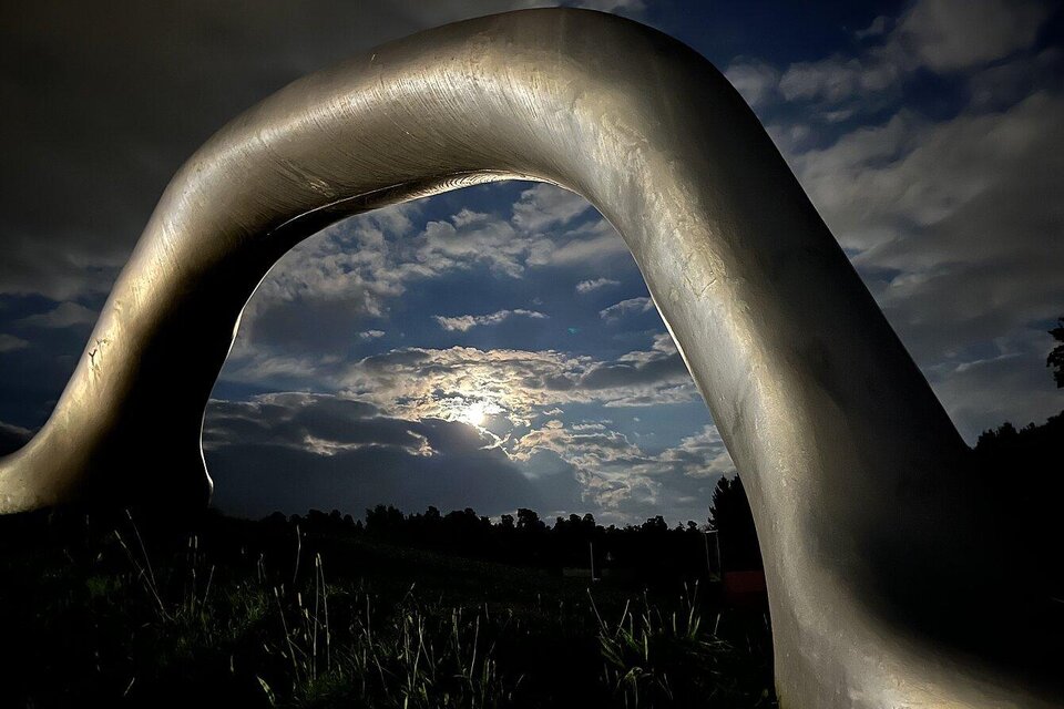 Eine geschwungene Metallskulptur steht vor einem bewölkten Himmel. Die Sonne scheint schwach durch die Wolken und verleiht der Szene eine mystische Stimmung. | © elisabeth fiedler