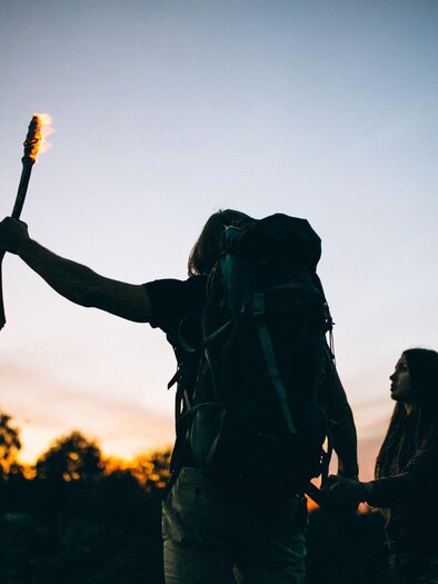 Two people with backpacks are standing outdoors at sunset. One person is holding a burning torch up high. | © Gemeinde Floing