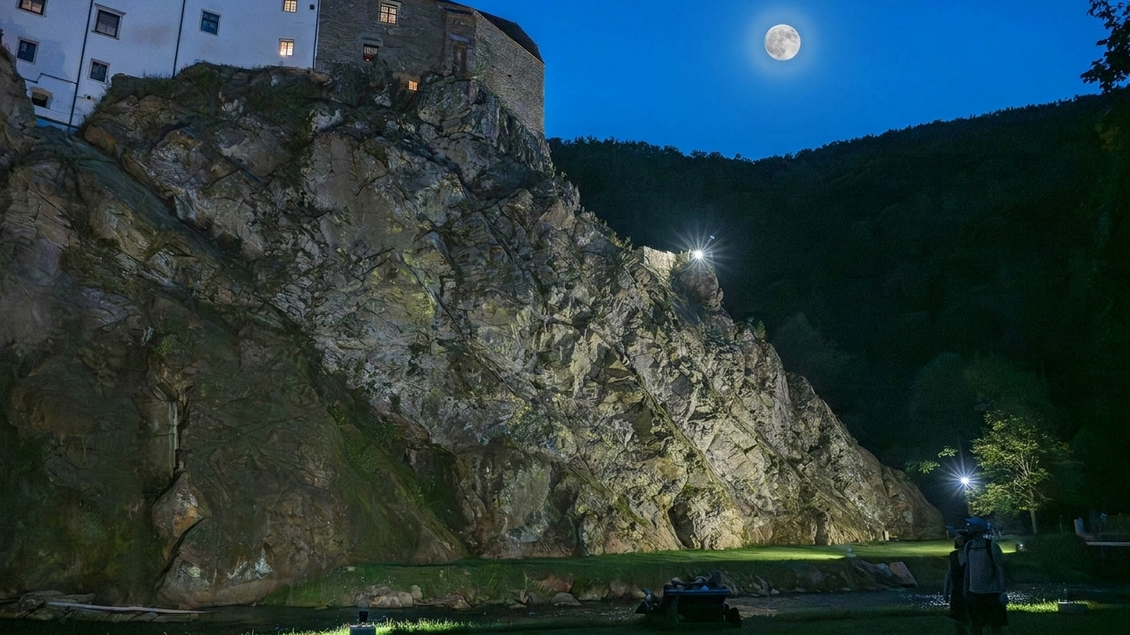 Eine steile Felswand bei Nacht, beleuchtet von Scheinwerfern. Im Hintergrund leuchtet ein großer, klarer Mond am Himmel. | © Gartenschloss Herberstein