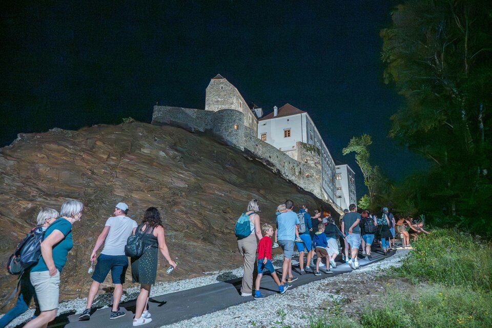 Eine Gruppe von Menschen geht auf einem Weg in der Nacht. Im Hintergrund ist eine Burg sichtbar, umgeben von Bäumen. | © Gartenschloss Herberstein