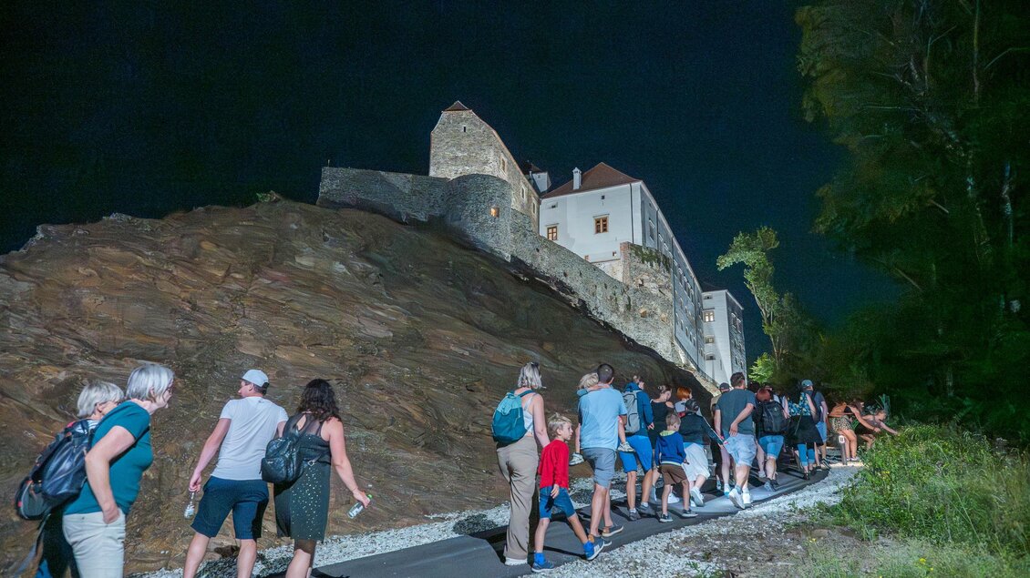 Eine Gruppe von Menschen geht auf einem Weg in der Nacht. Im Hintergrund ist eine Burg sichtbar, umgeben von Bäumen. | © Gartenschloss Herberstein