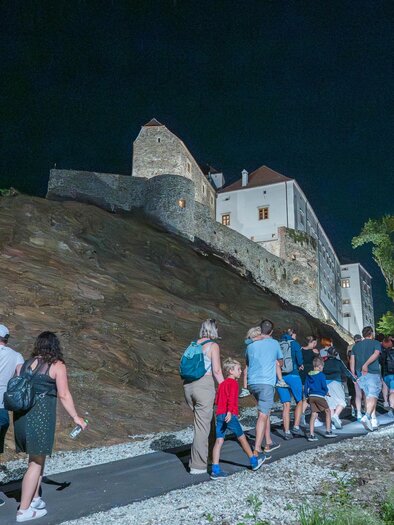 A group of people is walking on a path at night. In the background, a castle is visible, surrounded by trees. | © Gartenschloss Herberstein