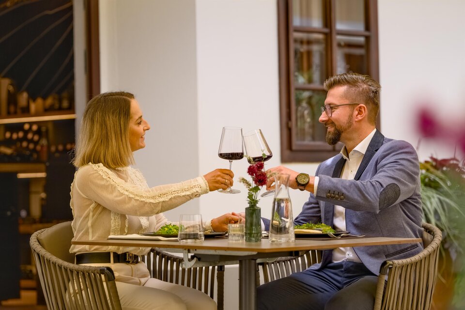 Ein Paar sitzt an einem Tisch und prostet sich mit Weingläsern zu. Auf dem Tisch sind Teller mit Essen und eine kleine Blume. | © Oststeiermark Tourismus