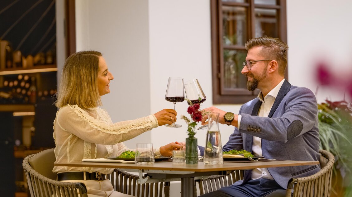 Ein Paar sitzt an einem Tisch und prostet sich mit Weingläsern zu. Auf dem Tisch sind Teller mit Essen und eine kleine Blume. | © Oststeiermark Tourismus