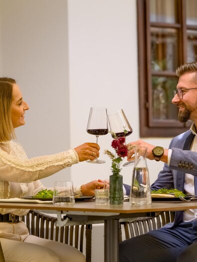 A couple sits at a table and toasts each other with wine glasses. On the table are plates with food and a small flower. | © Oststeiermark Tourismus