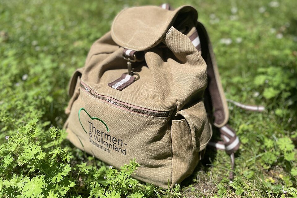 Ein beiger Rucksack liegt auf grünem Gras. Auf dem Rucksack ist das Logo der Thermen-Welt abgebildet. | © TV  Thermen- & Vulkanland