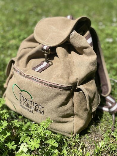 Ein beiger Rucksack liegt auf grünem Gras. Auf dem Rucksack ist das Logo der Thermen-Welt abgebildet. | © TV  Thermen- & Vulkanland