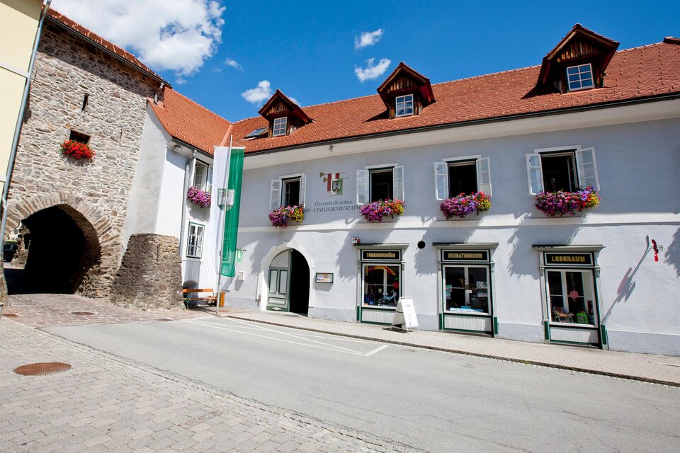 Ein historisches Gebäude mit bunten Blumenkästen und einem Tor in der Nähe. Der Himmel ist klar und blau, was eine angenehme Atmosphäre schafft.