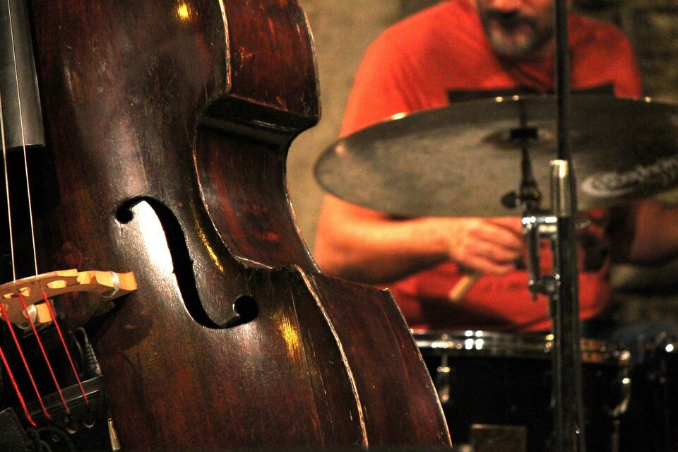 Ein Kontrabass steht im Vordergrund, während ein Schlagzeugspieler im Hintergrund spielt. Die Szenerie vermittelt eine lebendige musikalische Atmosphäre. | © Pixabay
