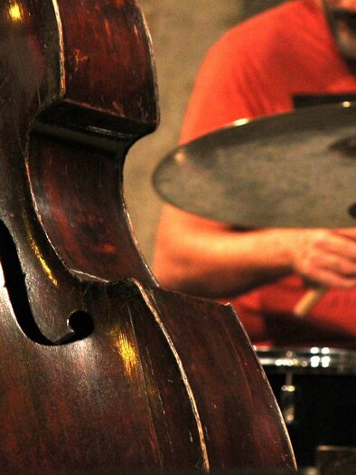 Ein Kontrabass steht im Vordergrund, während ein Schlagzeugspieler im Hintergrund spielt. Die Szenerie vermittelt eine lebendige musikalische Atmosphäre. | © Pixabay