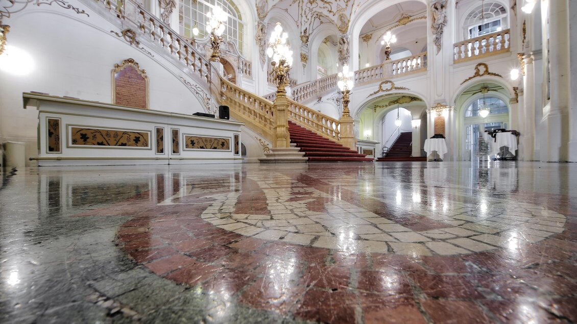 Ein prächtiger Innenraum mit einer großen Treppe und kunstvollen Verzierungen. Der polierte Boden reflektiert das Licht der eleganten Kronleuchter. | © (c) Opernhaus Graz -