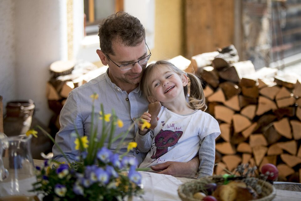 Ein fröhlicher Vater sitzt mit seiner lächelnden Tochter am Tisch. Im Hintergrund sind Holzscheite und frische Blumen zu sehen.