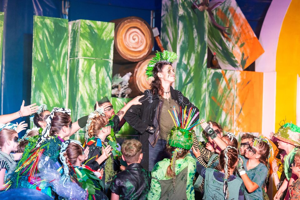 Eine lebendige Theateraufführung mit vielen Kindern in bunten Kostümen. Im Mittelpunkt steht eine Frau mit einem grünen Kopfputz, umgeben von begeisterten Darstellern.
