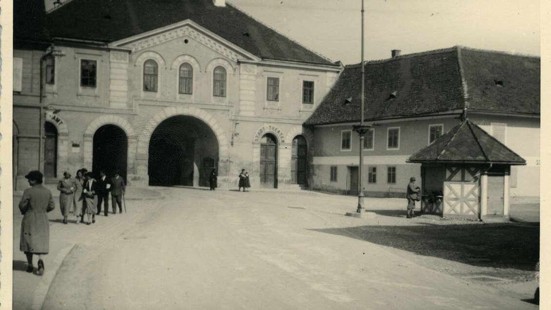 Historische Schwarz-Weiß-Aufnahme des Stadttors in Fürstenfeld, Steiermark, mit Passanten in zeittypischer Kleidung, markanten Torbögen und angrenzenden Gebäuden – Einblick in das frühere Stadtleben und kulturelles Erbe der Erlebnisregion Thermen- & Vulkanland. | © Museum Pfeilburg