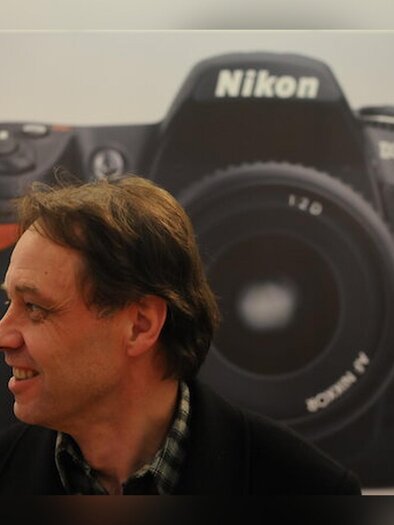 Speaker Rainer Fuchs in profile in front of a large-format depiction of a Nikon camera. | © Rainer Fuchs