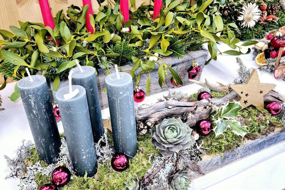 Ein festlicher Tischschmuck mit vier grauen Kerzen, umgeben von Moos und Weihnachtsdekoration. Rote Kugeln und grüne Zweige verleihen der Dekoration einen festlichen Charme. | © Sabine Fischer