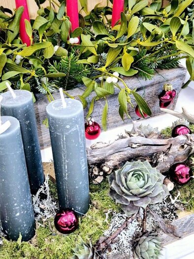 Ein festlicher Tischschmuck mit vier grauen Kerzen, umgeben von Moos und Weihnachtsdekoration. Rote Kugeln und grüne Zweige verleihen der Dekoration einen festlichen Charme. | © Sabine Fischer