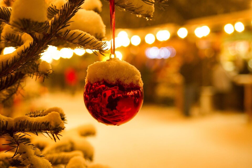Eine glänzende rote Weihnachtskugel hängt an einem schneebedeckten Baum. Im Hintergrund sind sanfte Lichter eines Märkte zu sehen. | © Stadtmarketing Bad Aussee