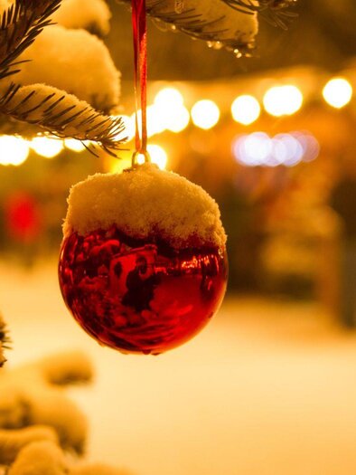 Eine glänzende rote Weihnachtskugel hängt an einem schneebedeckten Baum. Im Hintergrund sind sanfte Lichter eines Märkte zu sehen. | © Stadtmarketing Bad Aussee