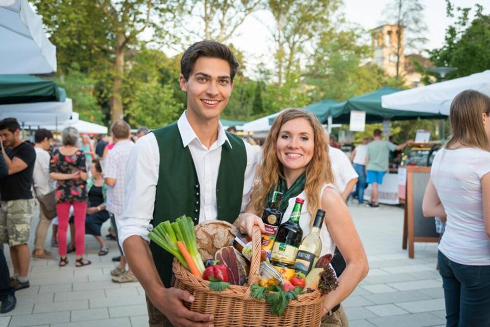 Eine junge Frau und ein junger Mann stehen auf einem Markt und halten einen Korb voller frischer Lebensmittel. Im Hintergrund sind weitere Marktstände und Menschen zu sehen. | © Vulkanlandmarkt Bad Gleichenberg