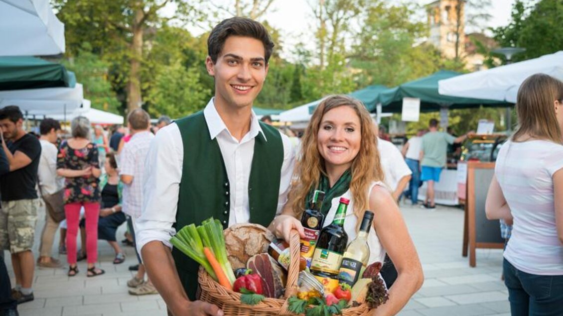 Eine junge Frau und ein junger Mann stehen auf einem Markt und halten einen Korb voller frischer Lebensmittel. Im Hintergrund sind weitere Marktstände und Menschen zu sehen. | © Vulkanlandmarkt Bad Gleichenberg