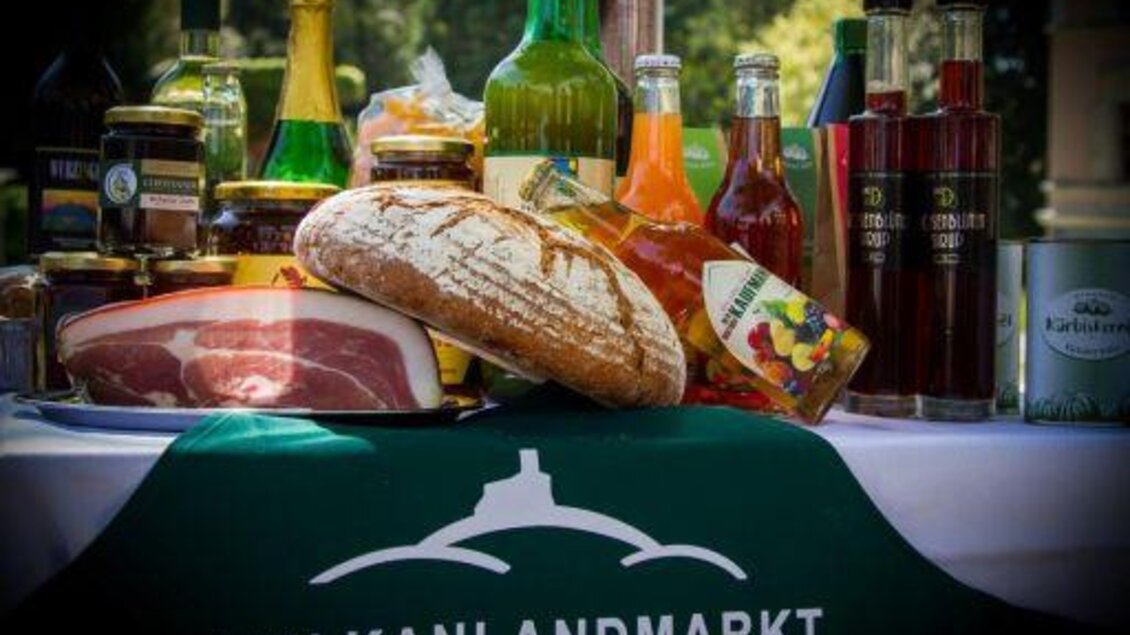 Ein Tisch voller regionaler Spezialitäten, darunter Brot, Schinken und verschiedene Getränke. Im Vordergrund ist das Logo des Vulkanlandmarktes Bad Gleichenberg sichtbar. | © Vulkanlandmarkt
