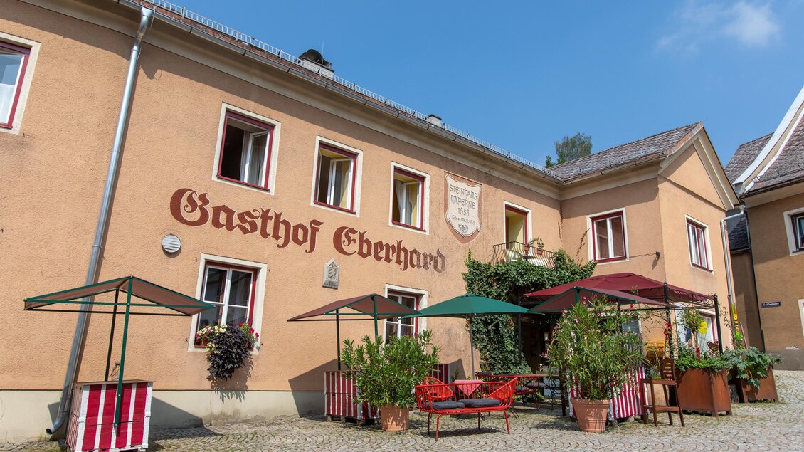 Ein einladender Gasthof mit roten und grünen Sonnenschirmen im Innenhof. Die Fassade ist in warmen Farben gehalten und mit Pflanzen dekoriert. | © Ilse Blachfellner-Mohri