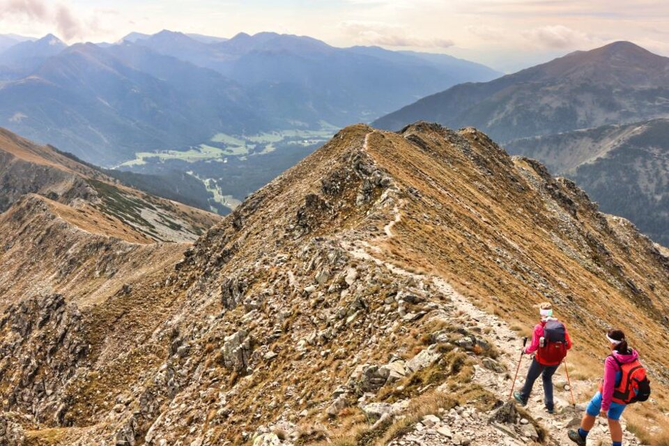 Zwei Wanderer stehen auf einem schmalen Bergweg mit beeindruckendem Ausblick auf die umliegenden Berge und das Tal. Die Landschaft ist wild und schön, umgeben von goldbraunem Gras und Felsen. | © WEGES OG