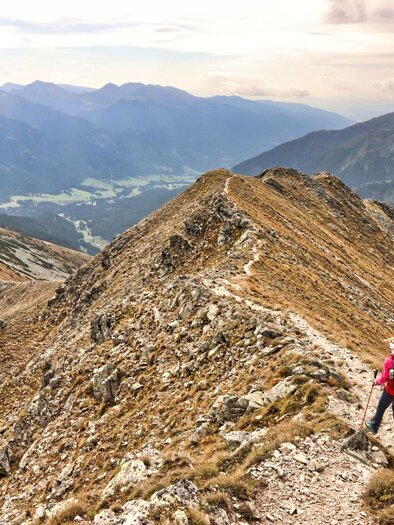 Zwei Wanderer stehen auf einem schmalen Bergweg mit beeindruckendem Ausblick auf die umliegenden Berge und das Tal. Die Landschaft ist wild und schön, umgeben von goldbraunem Gras und Felsen. | © WEGES OG