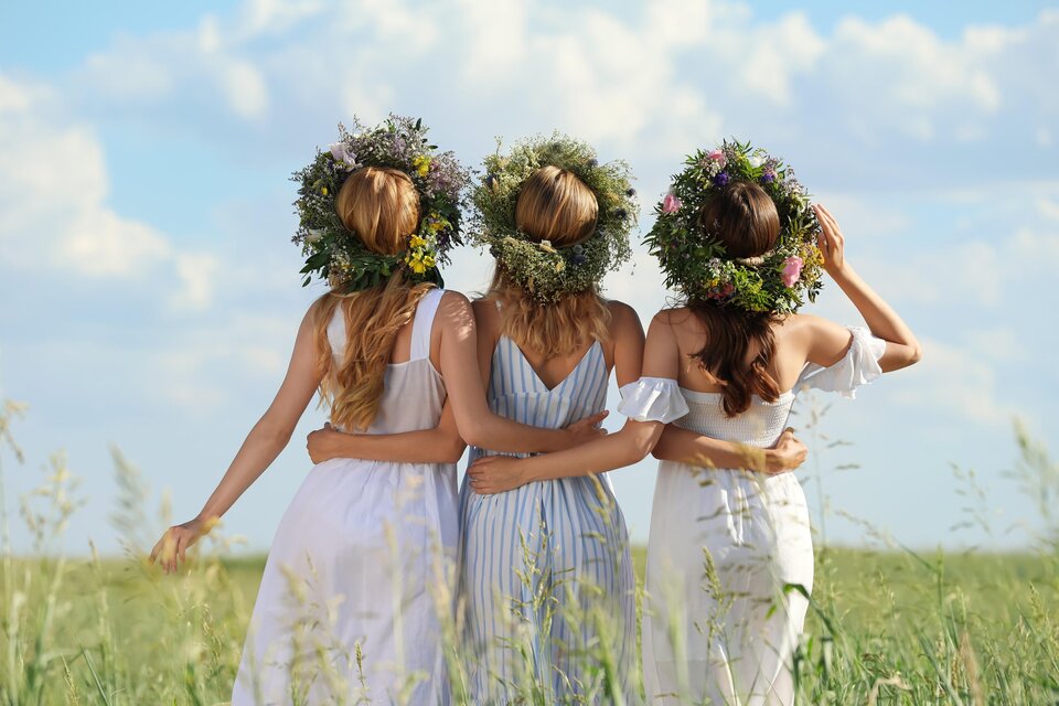 Drei Frauen in hellen Sommerkleidern stehen in einem Feld. Sie tragen Blumenkränze auf ihren Köpfen und blicken in die Ferne. | © Loibner