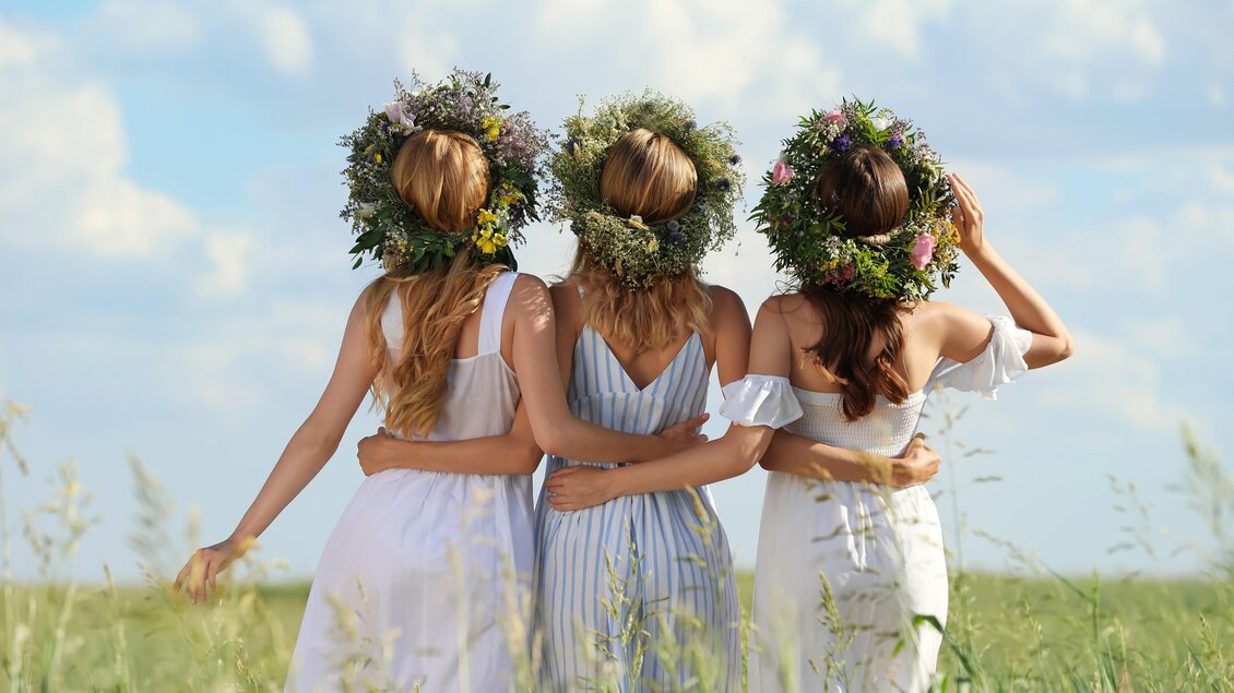 Drei Frauen in hellen Sommerkleidern stehen in einem Feld. Sie tragen Blumenkränze auf ihren Köpfen und blicken in die Ferne. | © Loibner
