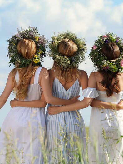 Drei Frauen in hellen Sommerkleidern stehen in einem Feld. Sie tragen Blumenkränze auf ihren Köpfen und blicken in die Ferne. | © Loibner