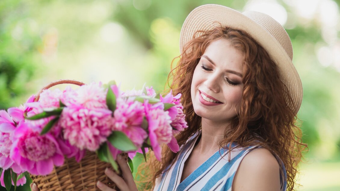 Eine lächelnde Frau mit lockigem Haar trägt einen Strohhut und hält einen Korb voller rosafarbener Blumen. Im Hintergrund sind grüne Bäume und eine sonnige Umgebung zu sehen. | © Loibner