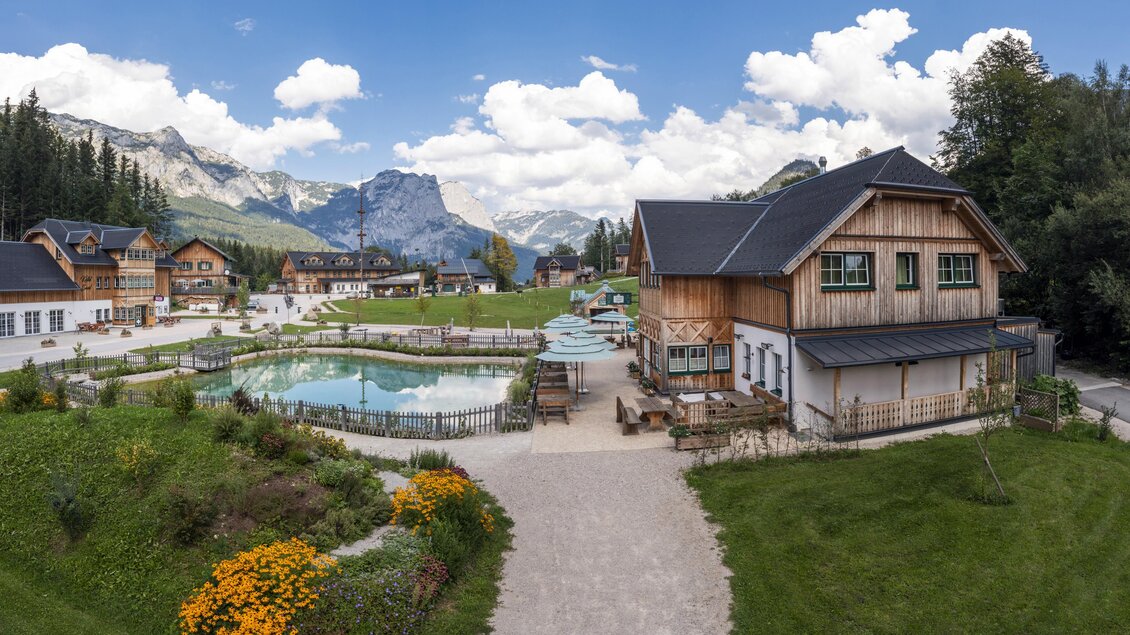 Eine malerische Landschaft mit einem Holzgebäude und einem blauen Pool. Im Hintergrund sind hohe Berge und eine grüne Wiese zu sehen. | © www.zloam.at