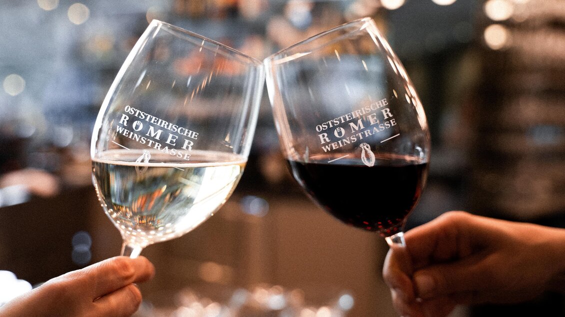 Zwei Weingläser stoßen an, eines mit Weißwein und eines mit Rotwein. Im Hintergrund ist eine stilvolle Atmosphäre mit unscharfen Lichtern zu sehen. | © (c)Heilthermen Resort Bad Waltersdorf