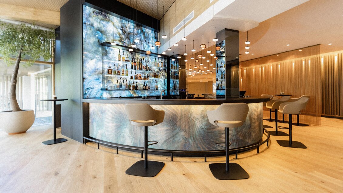 Eine elegante Bar mit einem skulpturalen Bartresen und modernen Stühlen. Im Hintergrund ist eine ansprechende Barbeleuchtung und eine dekorative Wand zu sehen. | © Heiltherme Bad Waltersdorf_Eisenberger