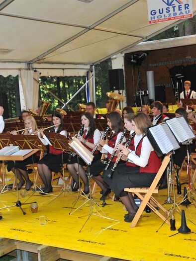 A wind orchestra plays under a tent. The musicians wear red vests and enjoy the performance. | © Walfest_Archiv TVB
