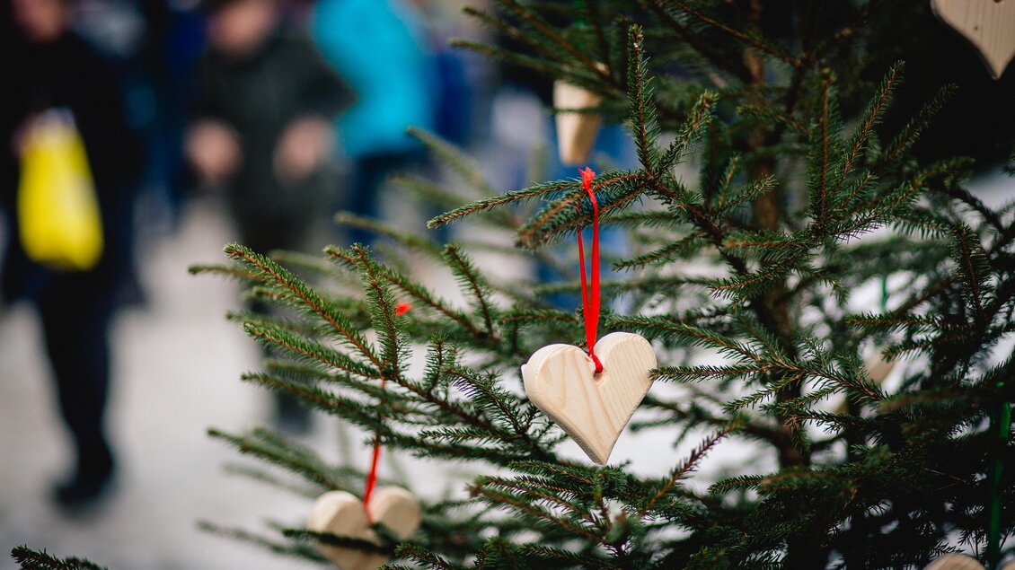 Ein mit Holzherzen geschmückter Weihnachtsbaum steht im Vordergrund. Im Hintergrund sind unscharfe Menschen zu sehen, die sich bewegen. | © TV Hochsteiermark, nicoleseiser.at