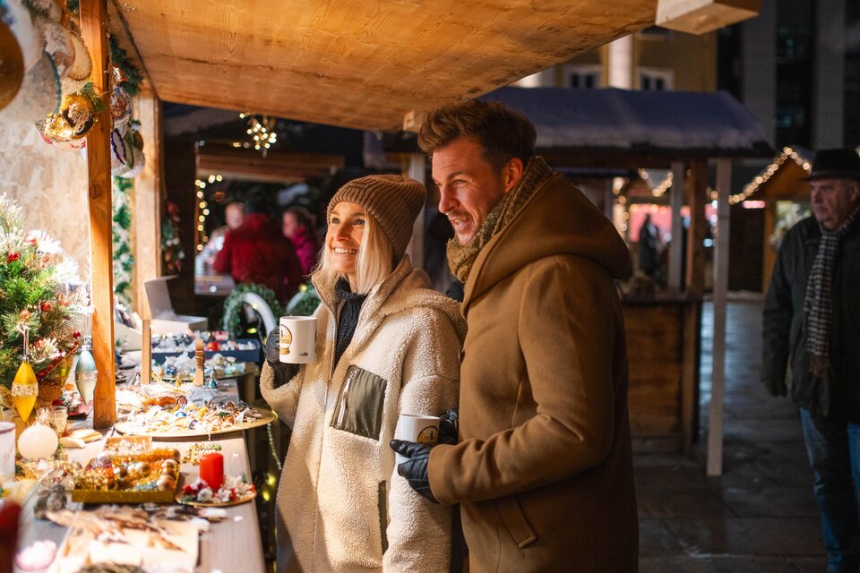 Ein festlicher Weihnachtsmarkt mit leuchtenden Ständen und fröhlichen Menschen. Ein Paar genießt heiße Getränke und betrachtet die angebotenen Waren. | © Ekatarina Paller
