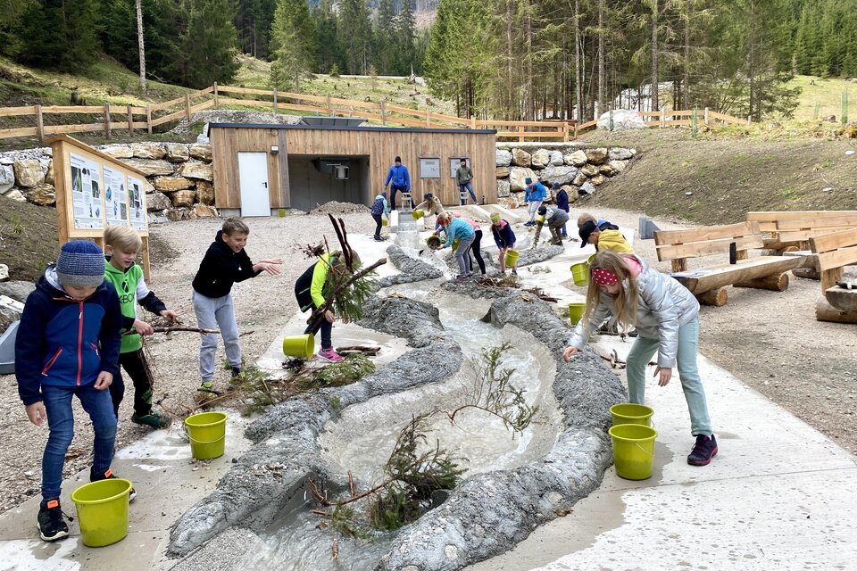 Eine Gruppe von Kindern spielt draußen und arbeitet mit Wasser an einem kleinen Flussbett. Im Hintergrund sind Wälder und Berge zu sehen. | © Wassererlebnis Öblarn